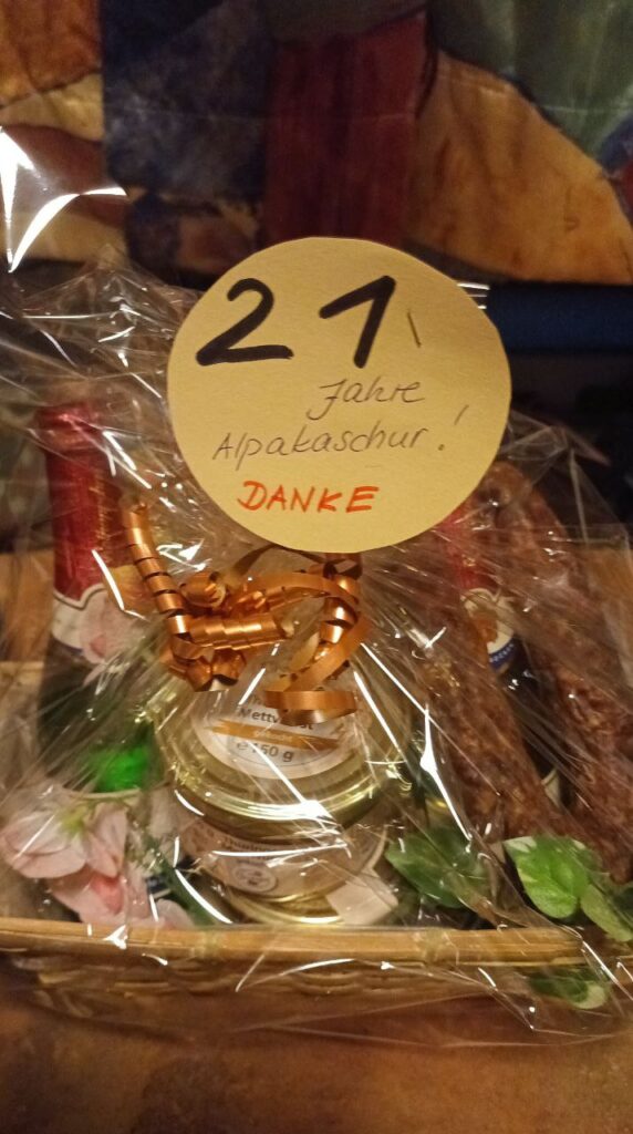 Ein Geschenk in einer durchsichtigen Verpackung, das eine handgeschriebene Note mit "21 Jahre Alpakaschur! DANKE" enthält, sowie verschiedene Essensartikel und Dekorationen.
